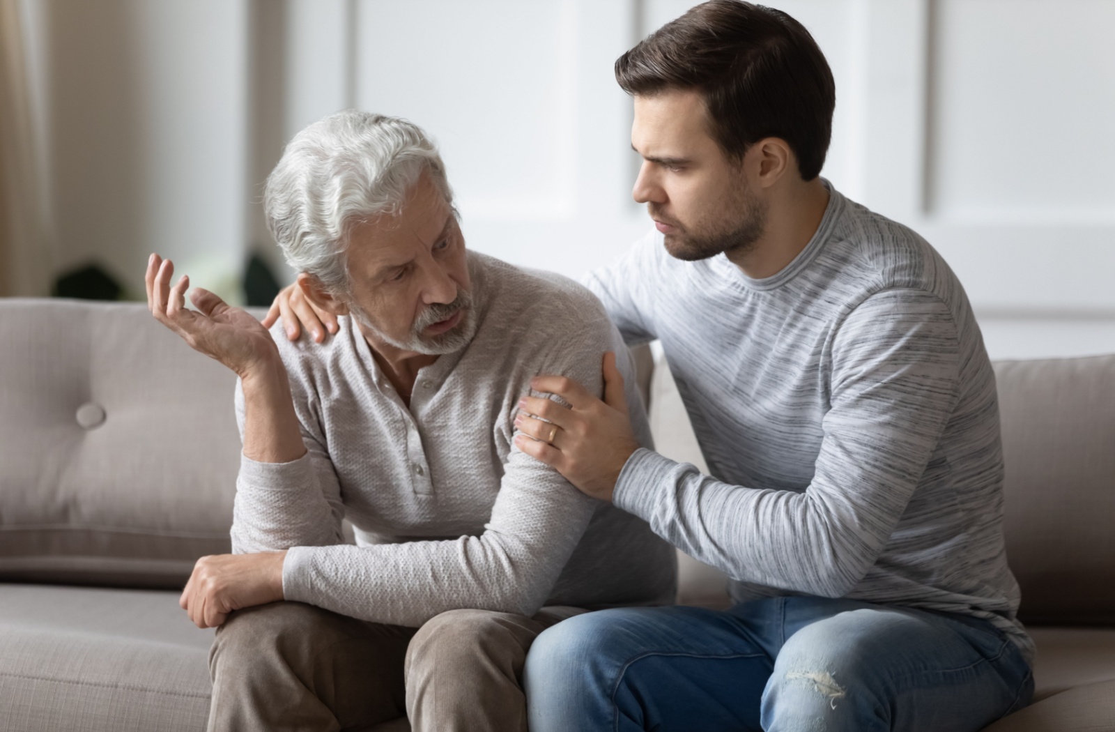 A young man comforting his senior father on the couch while discussing dementia.