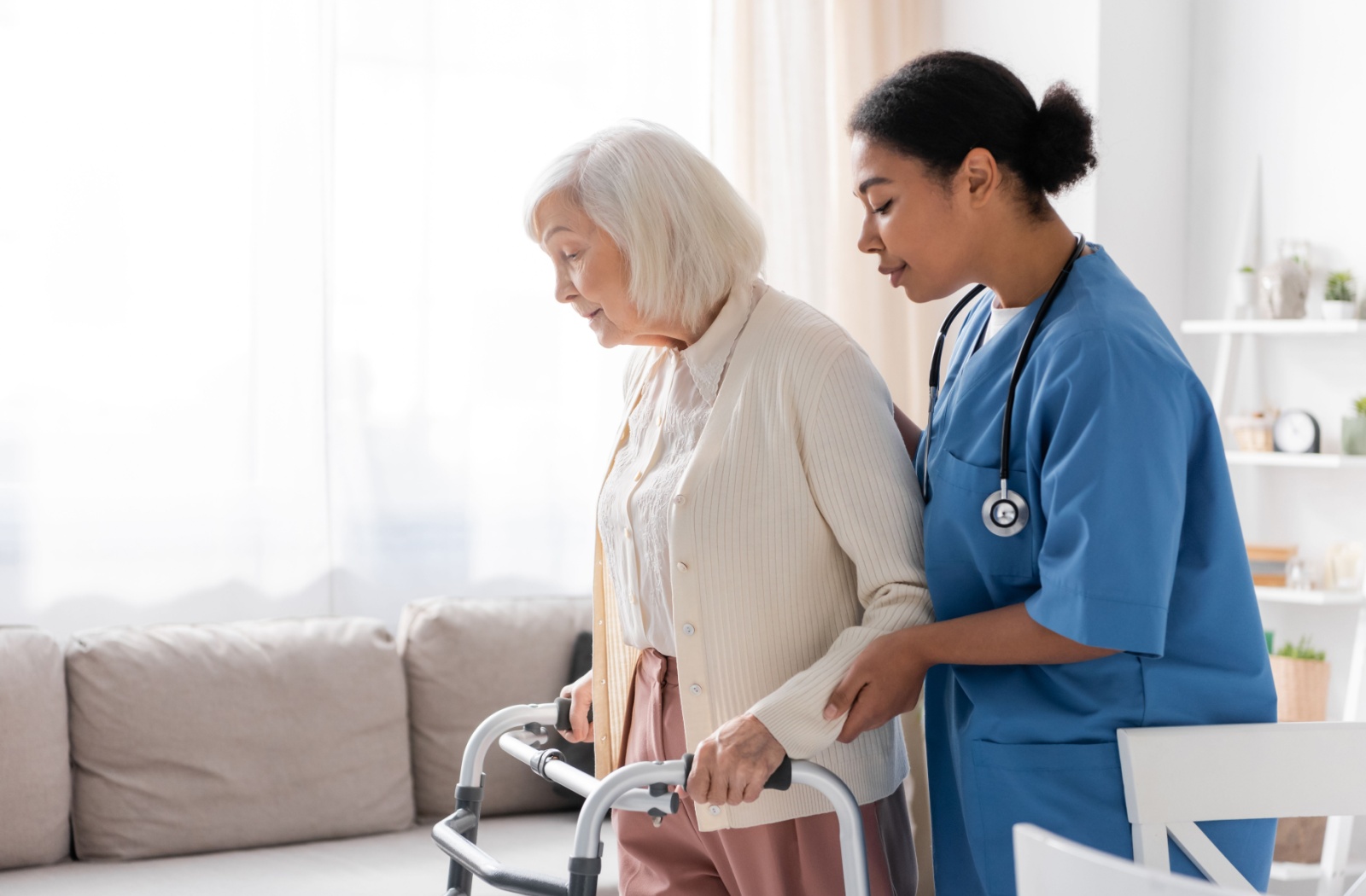 A skilled nurse helps an assisted living resident move using her walker in a brightly lit suite.