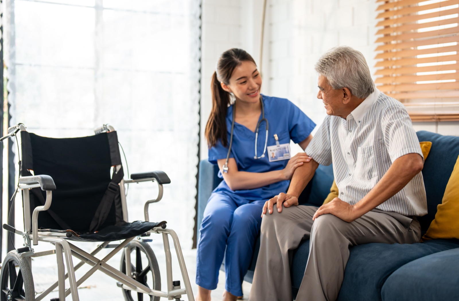 A caregiver and a senior man with mobility problems sitting on the couch smiling at one another getting ready to move him to a wheelchair.