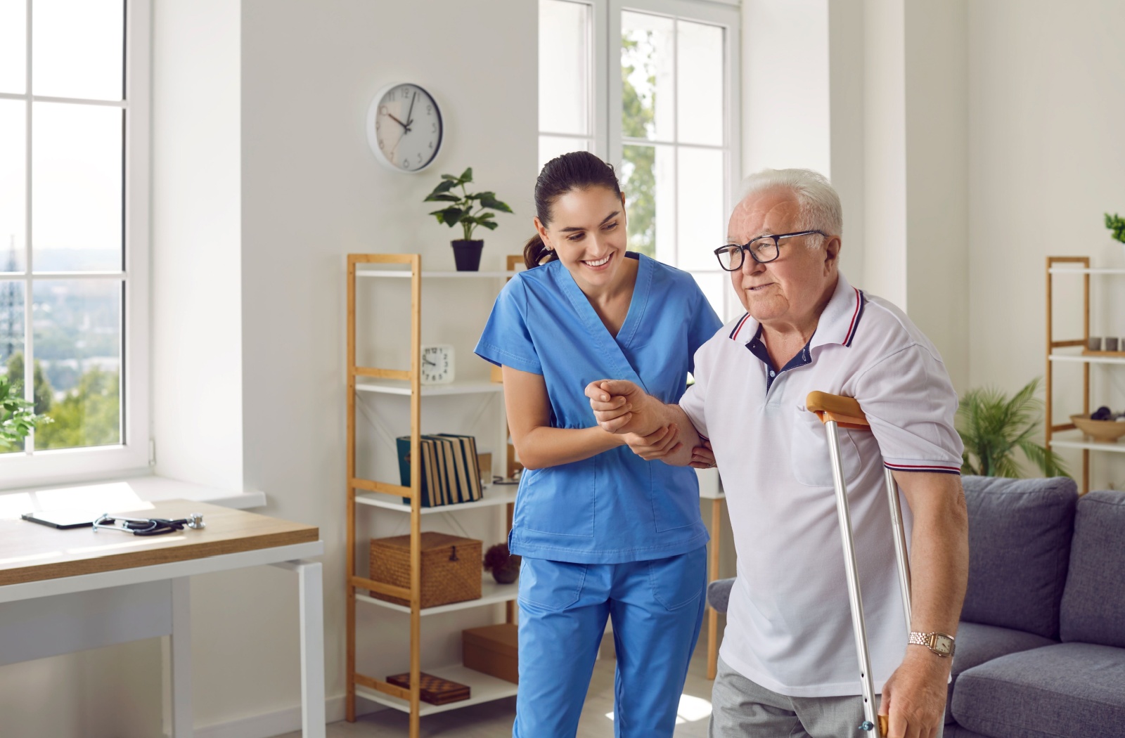 A caregiver holding a smiling senior man's arm to support him while he uses a crutch to walk again.
