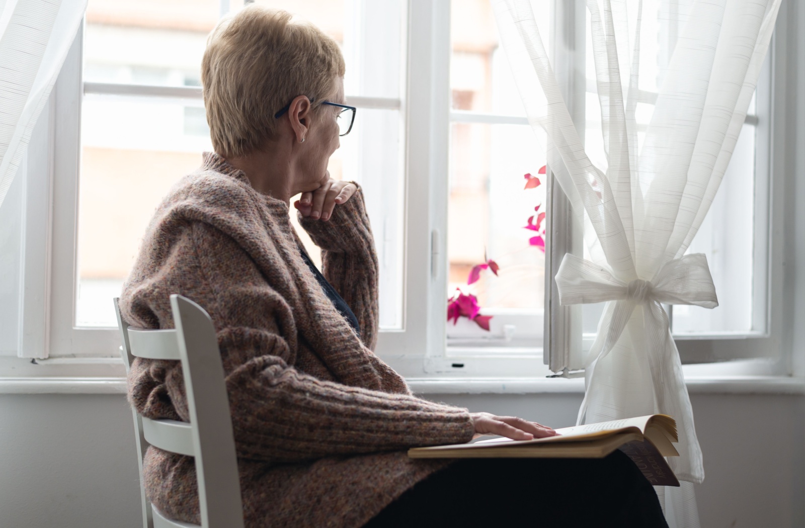 A senior sits on a chair with a book, looking thoughtfully out the open window beside them.