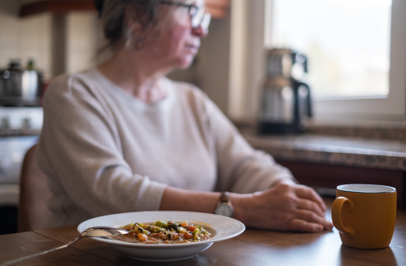 An older adult looking away from a bowl of soup on the table in front of them.