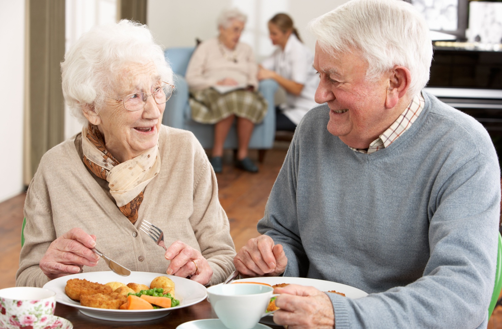 Two older adults enjoy a meal together in a senior living community while a nurse reads with another resident behind them.