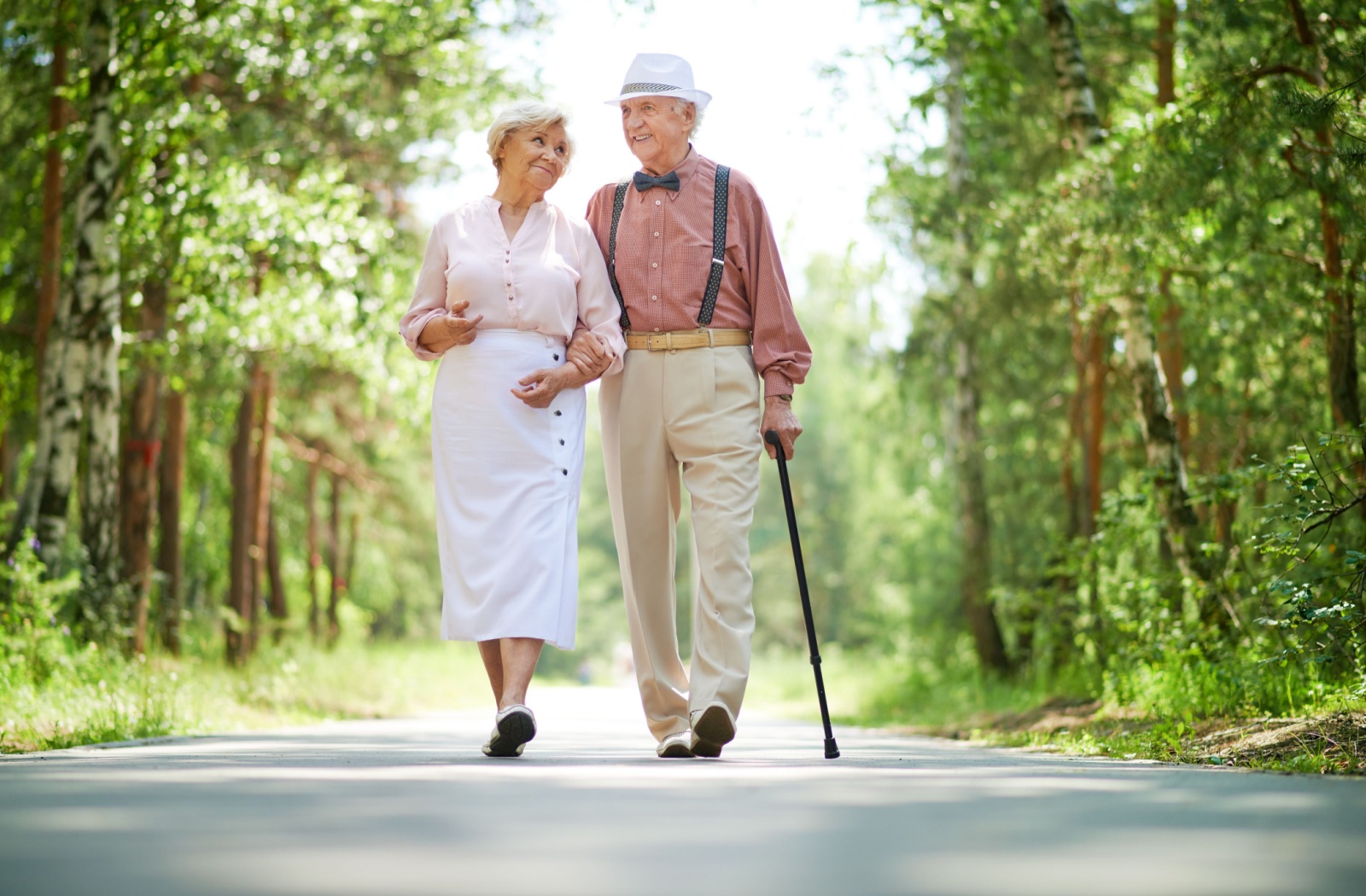 Senior man and woman walking on a path between woods. Man has a bowler hat, cane, bowtie, and suspenders.
