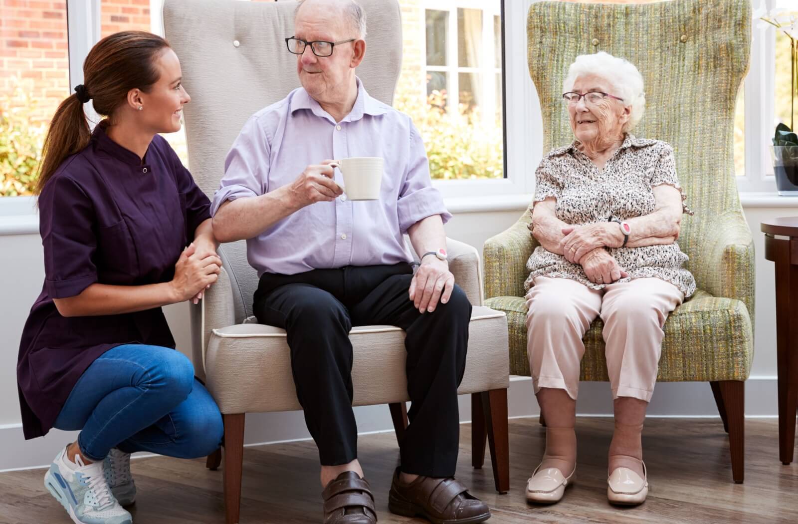 Senior man and woman sitting in armchairs with younger female caregiver kneeling next to man, who is holding a mug.