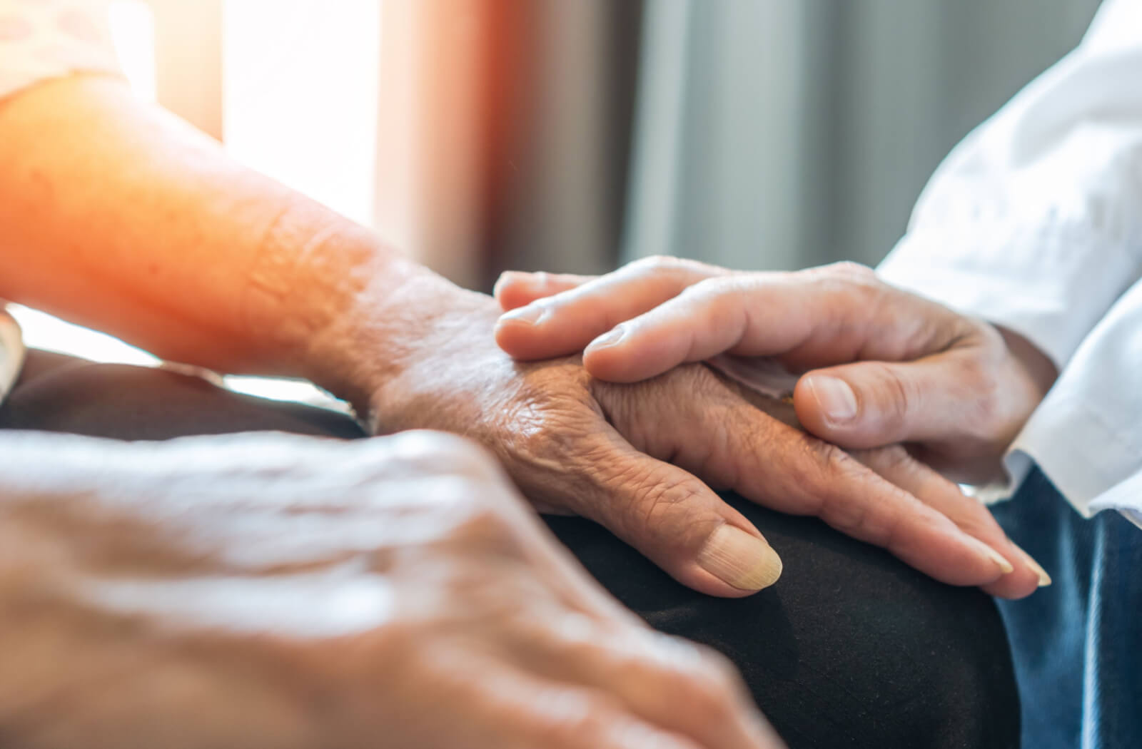 An older adult with Parkinson's disease sits in front of a sunlit window while an out-of-frame caregiver touches their hand to support them