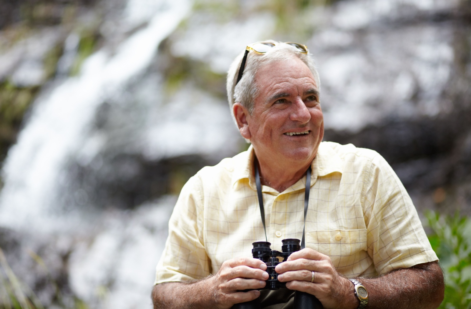 A senior holds a pair of binoculars in front of a waterfall, trying birdwatching for the first time.