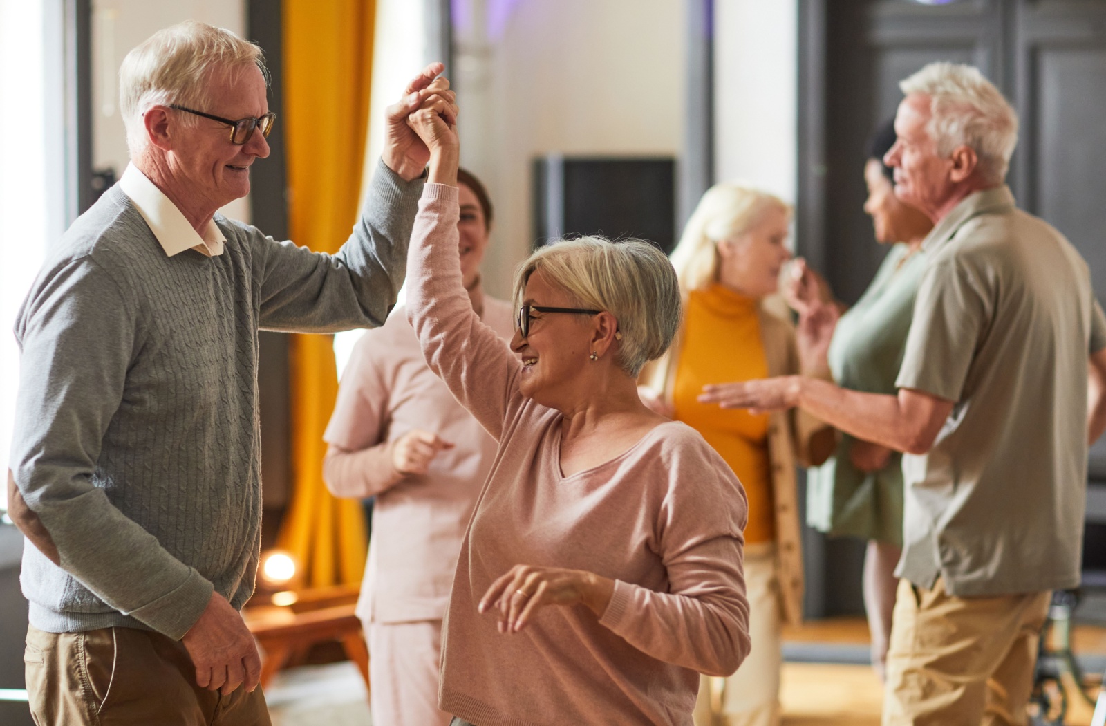 A group of seniors in a senior community practice line dance moves as a caregiver assists in the background.