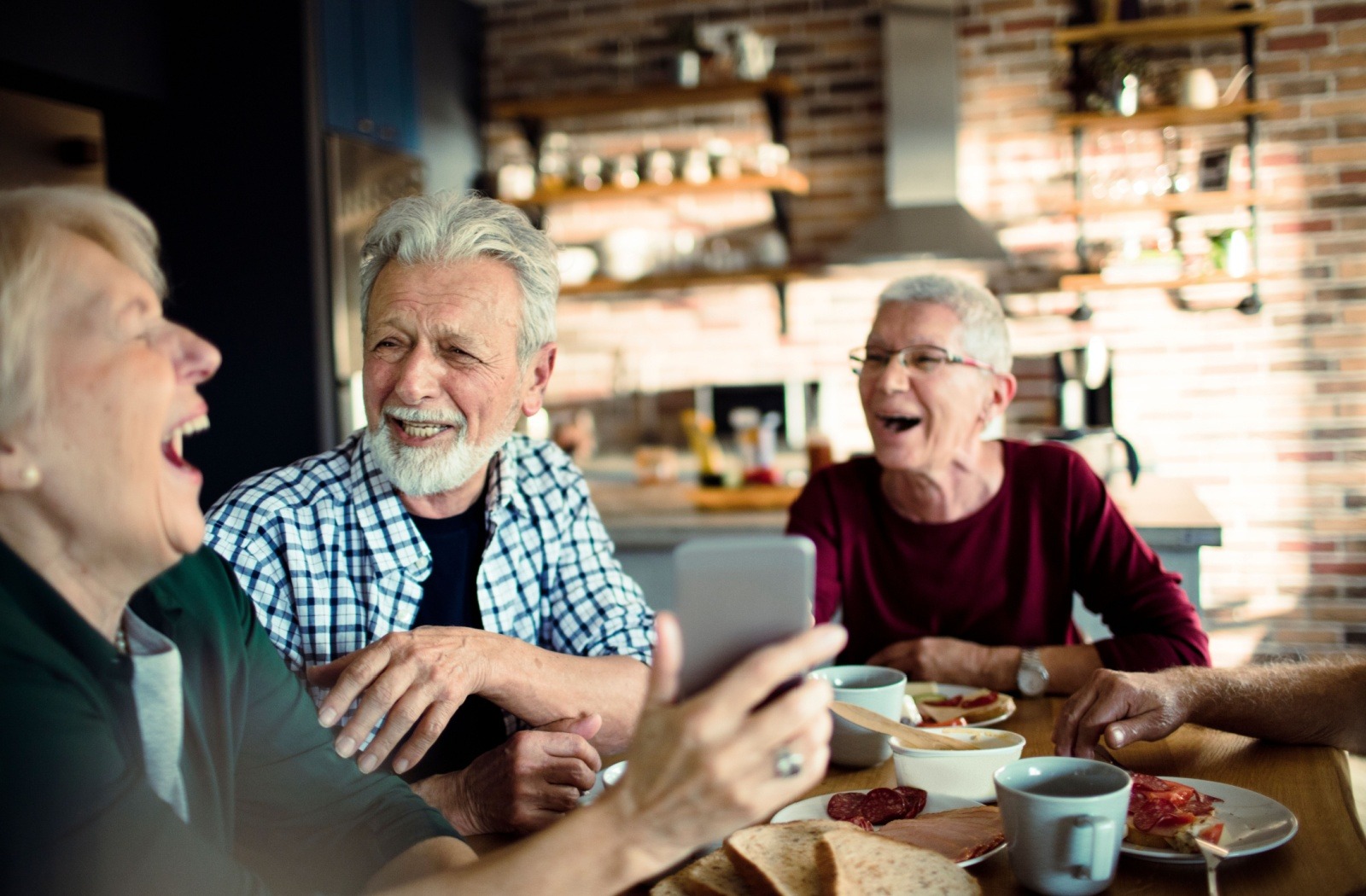 A group of senior friends laugh together while eating lunch and looking at something on a phone
